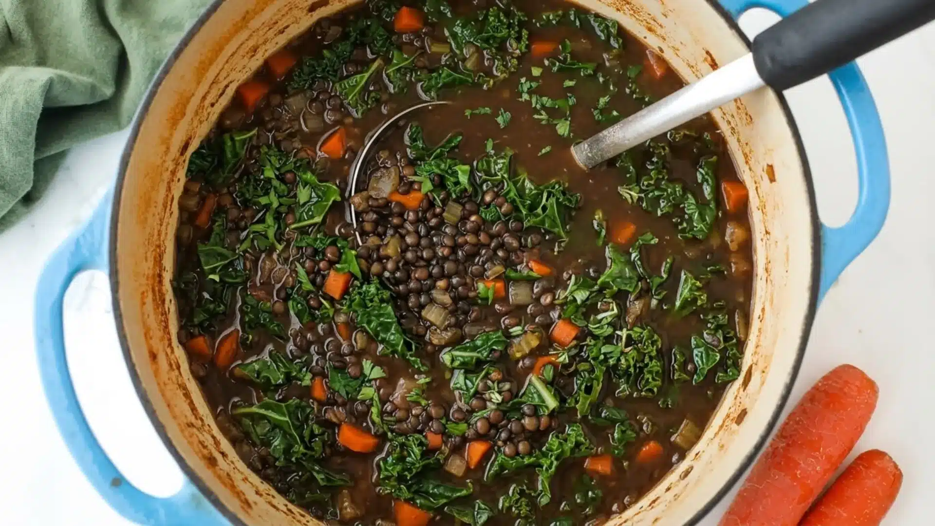 A blue enameled pot filled with hearty lentil soup featuring kale and carrots, with a ladle resting inside.