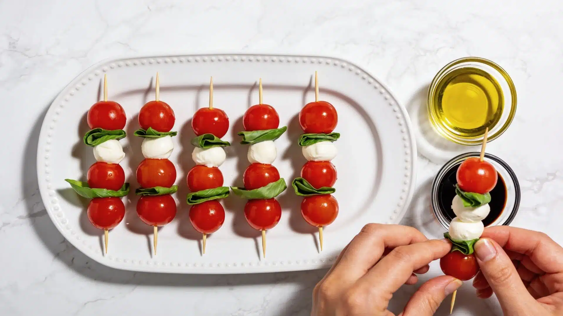 Caprese salad skewers with cherry tomatoes, mozzarella balls, and basil leaves arranged on a white platter, with a hand assembling one skewer over a bowl of balsamic glaze.