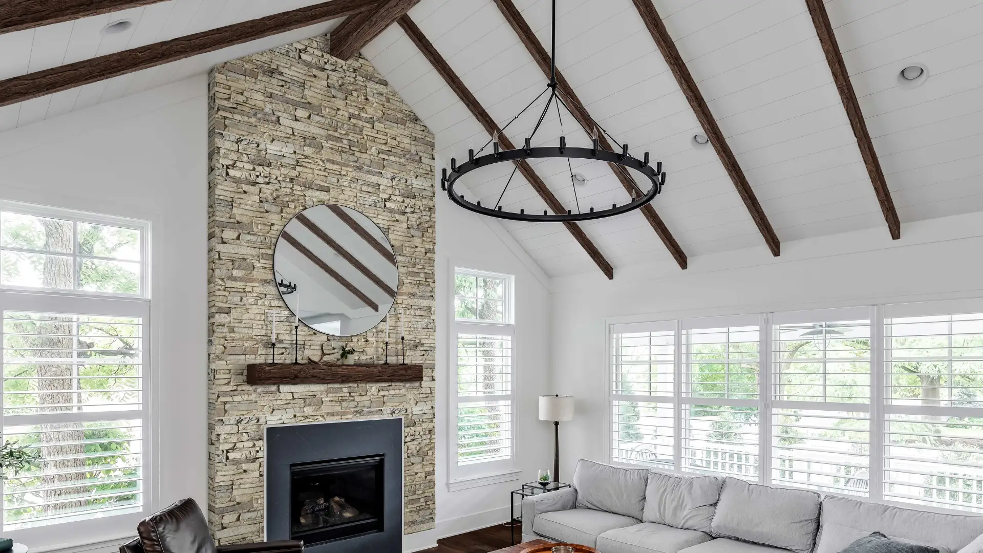 Vaulted ceiling living room with stone fireplace, large circular mirror, black chandelier, and white shuttered windows