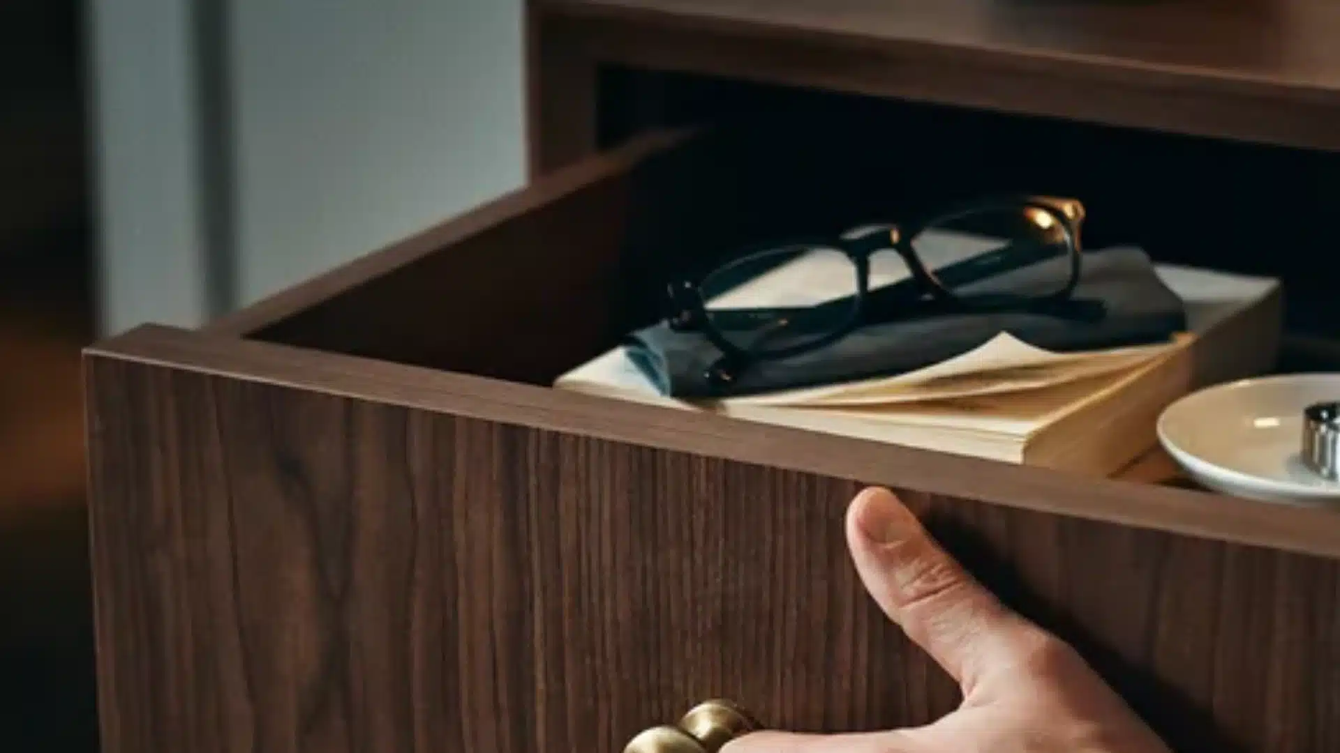 Hand opening a wooden drawer with glasses and papers inside, showing smooth metal slides on a nightstand.