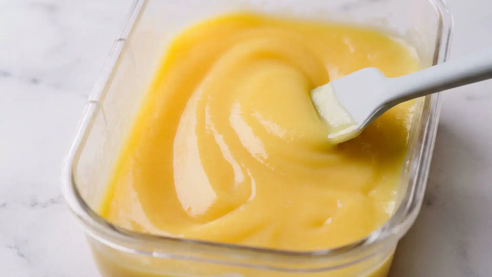homemade lemon curd in a glass jar with fresh lemons on a marble surface