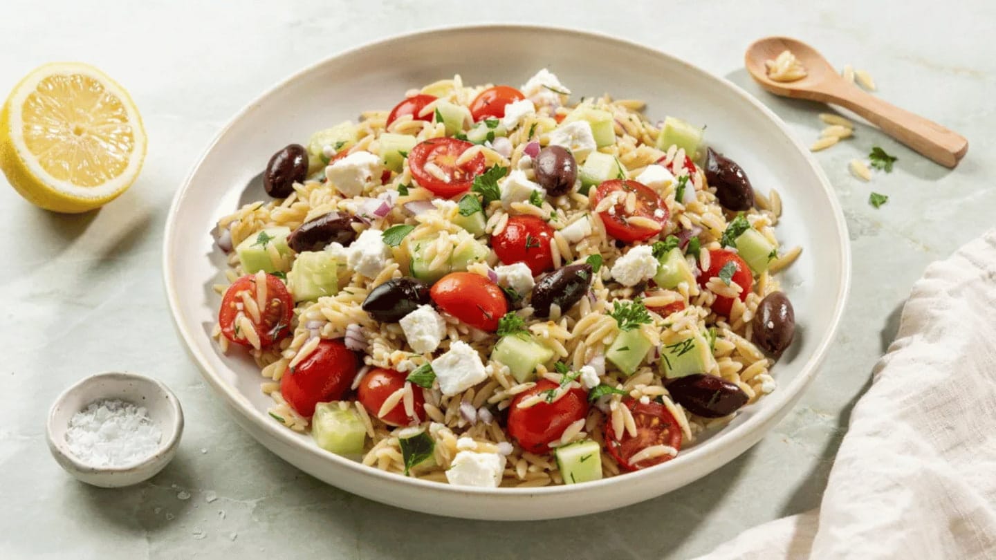 orzo salad with feta cheese, cherry tomatoes, cucumber, olives, and herbs served in a white bowl