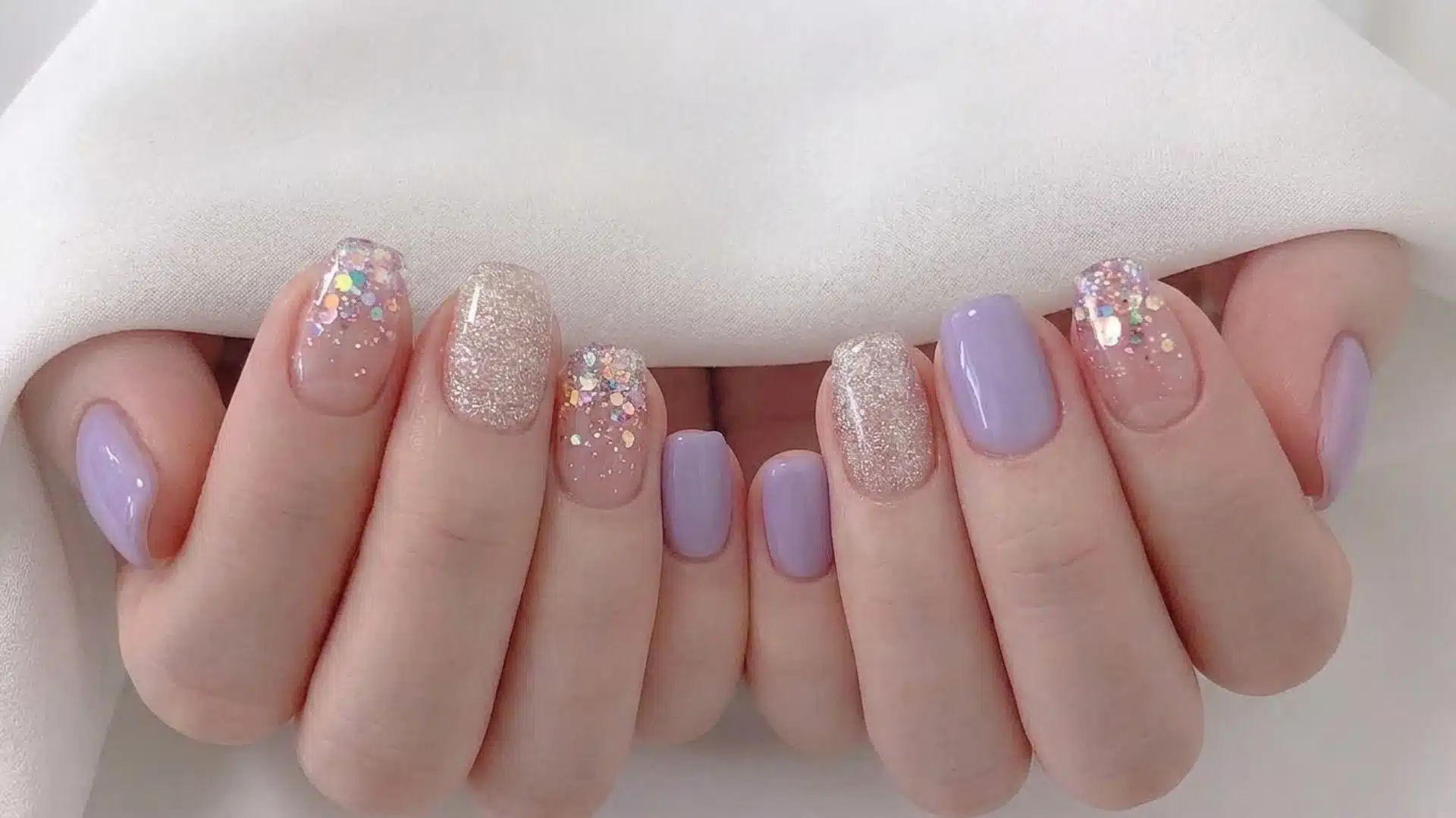 Hands display a variety of short, square-shaped manicures featuring lilac, glitter, and confetti nail polish designs.