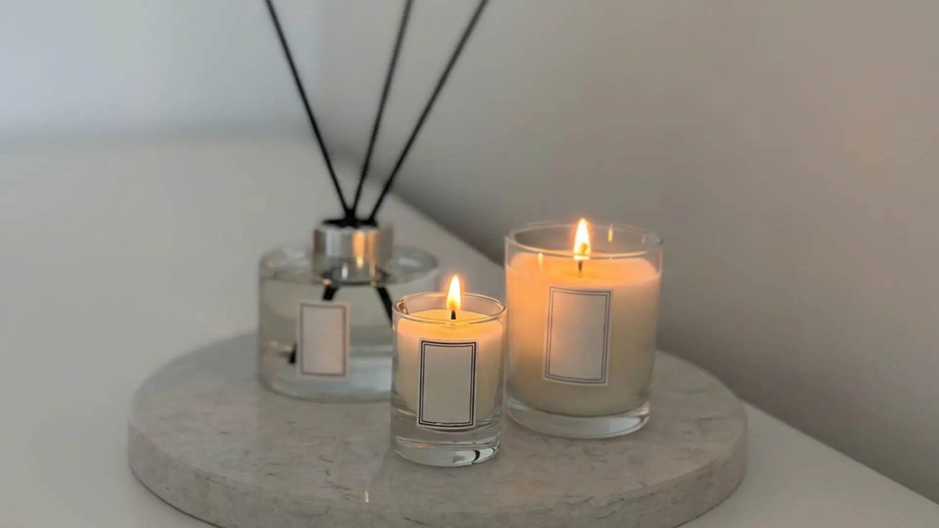 two lit soy candles in glass vessels and a reed diffuser on a marble tray