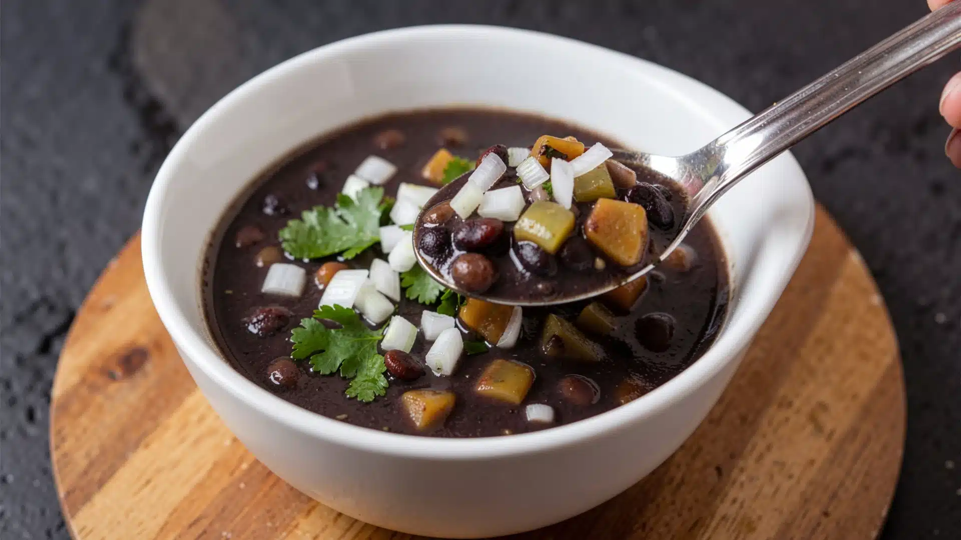 A spoon lifts a serving of black bean soup with diced vegetables and cilantro from a white bowl.