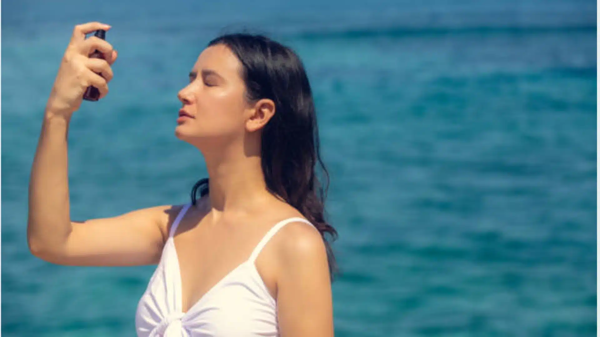Woman applying makeup setting spray outdoors by the ocean with eyes closed and natural sunlight glow