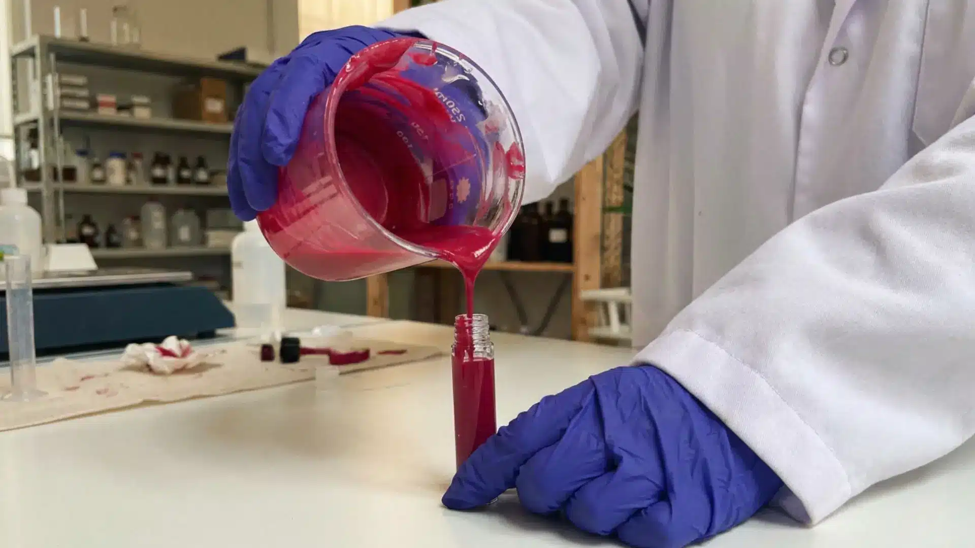 cosmetic lab technician in blue gloves pouring pink lip gloss formula into a tube
