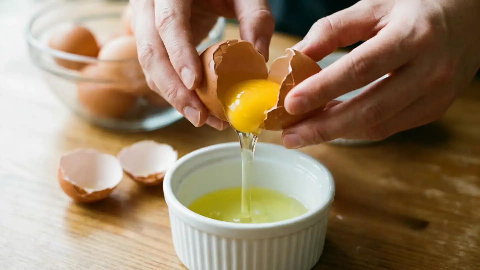 hands separating egg yolk from white into a white bowl with cracked eggshells on a wooden surface