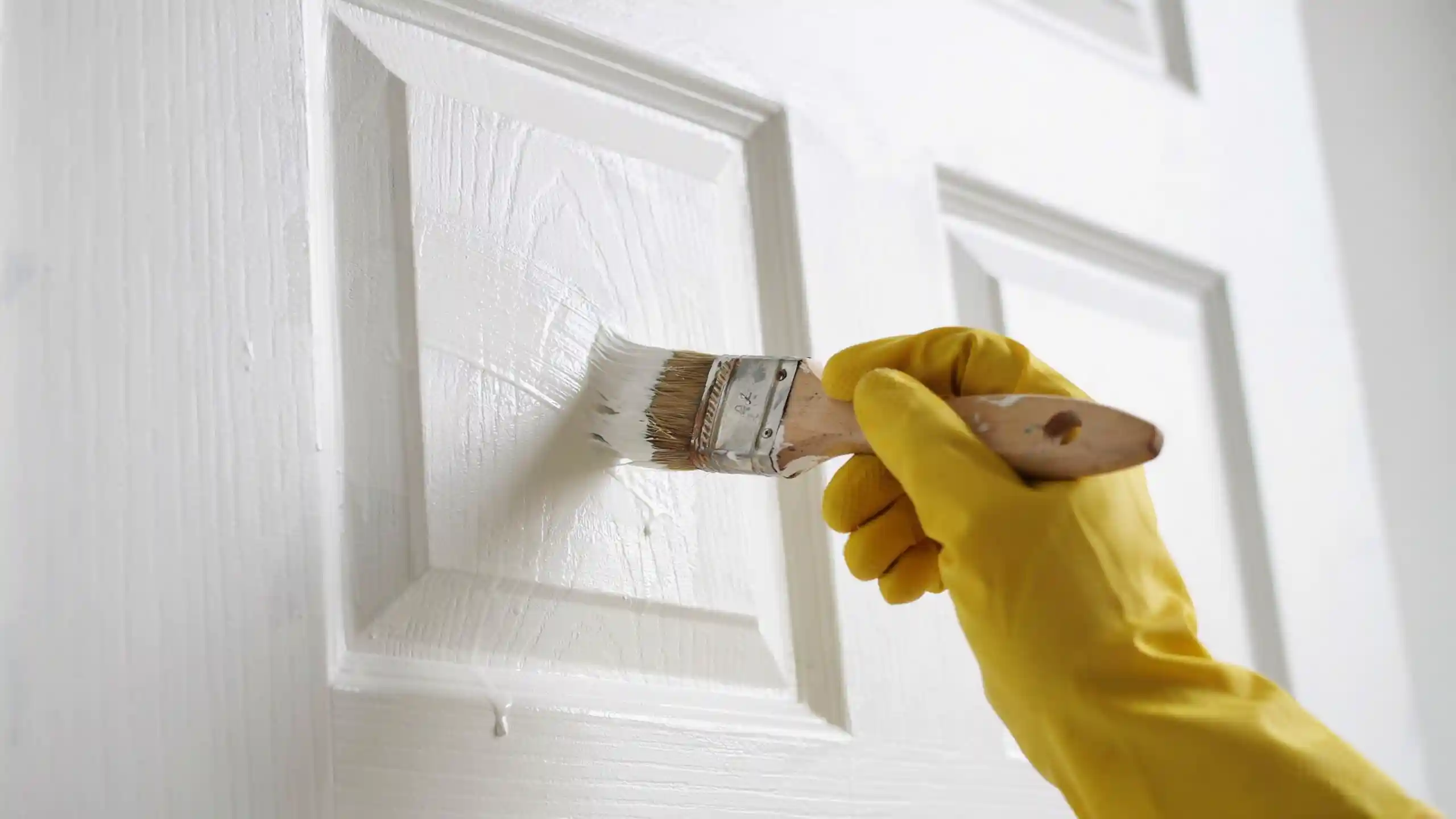 A hand wearing a yellow glove paints a white door with a brush.