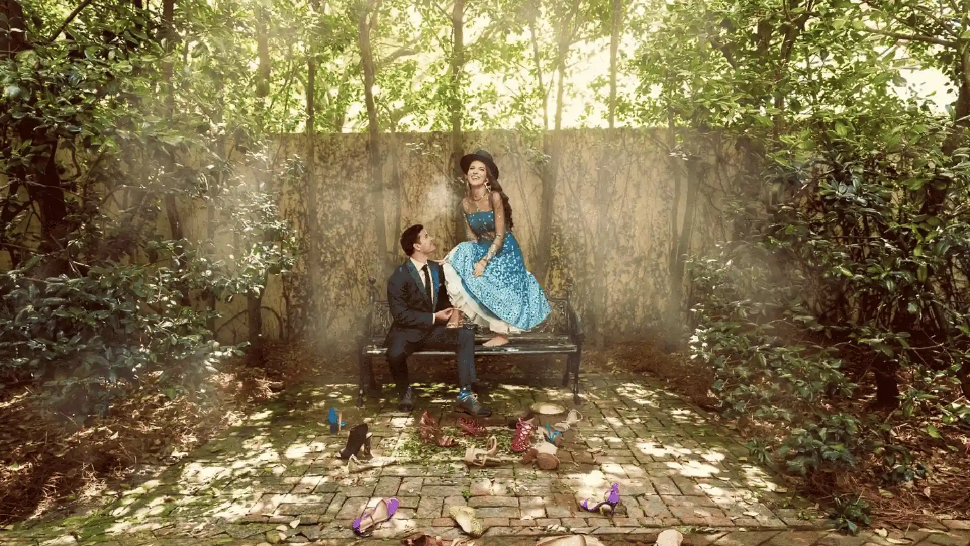 A man in a suit and a woman in a blue dress sit on a bench in a sun-dappled garden surrounded by greenery, with many pairs of shoes scattered on the brick path.