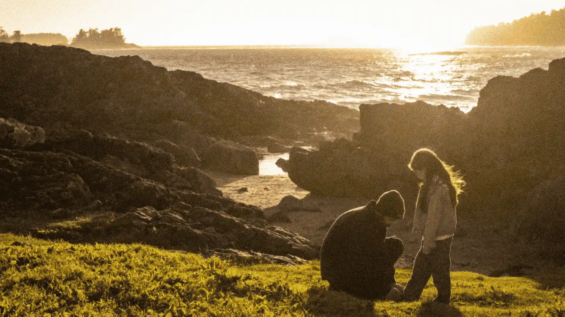 A person and a child crouch on a grassy knoll overlooking a rocky coastline and the ocean at sunset.