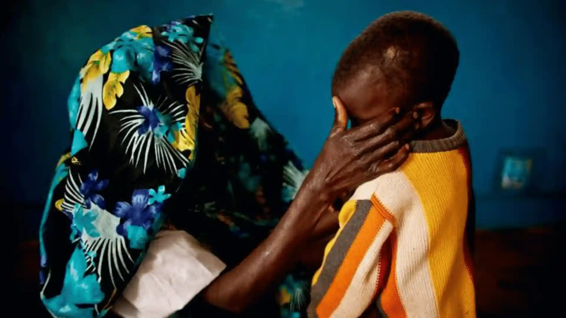 A person in a vibrant floral shirt holds a child in a striped sweater, with the child covering their face.