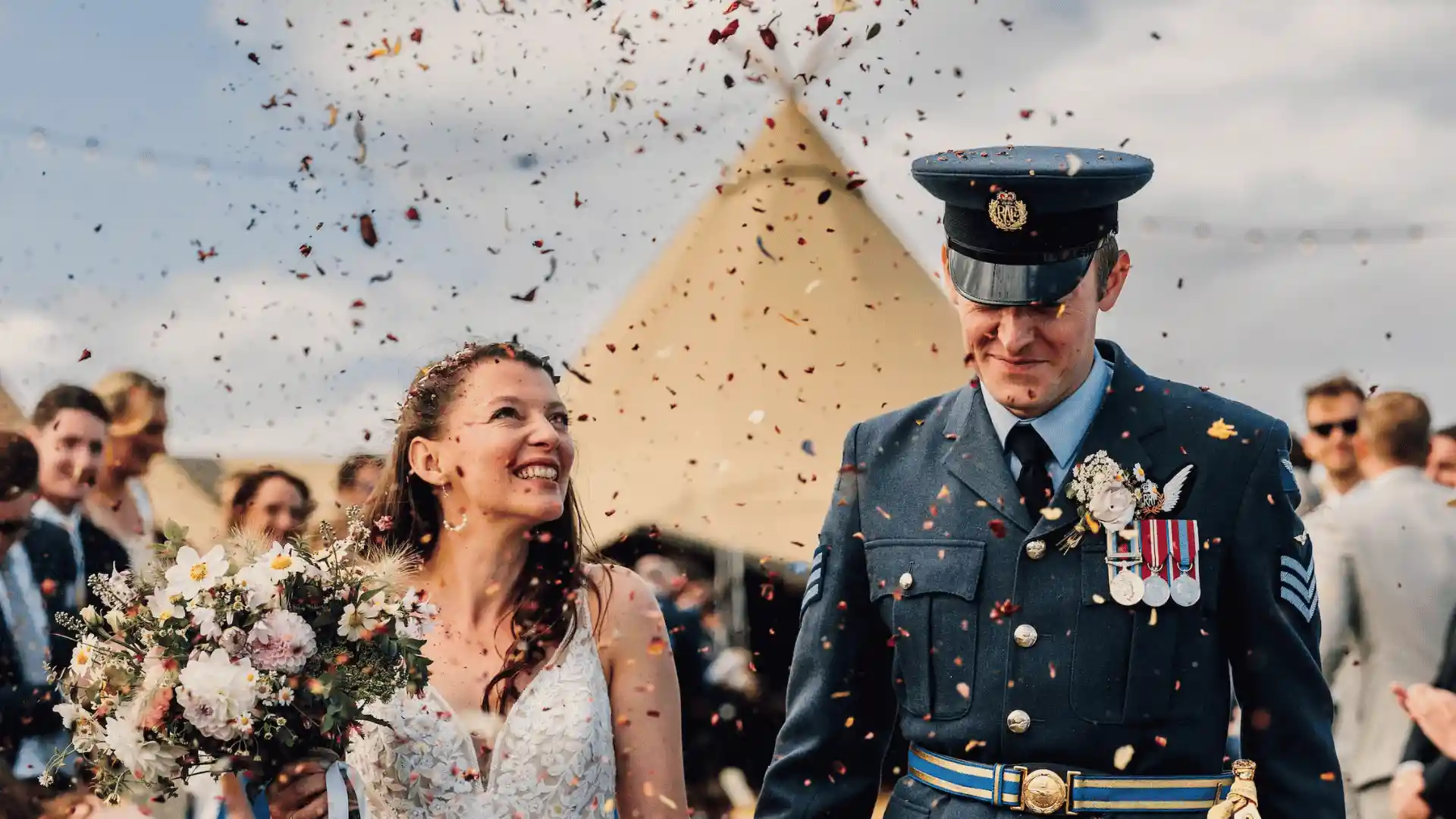 A smiling bride with a bouquet and a groom in a military uniform walk as confetti rains down.