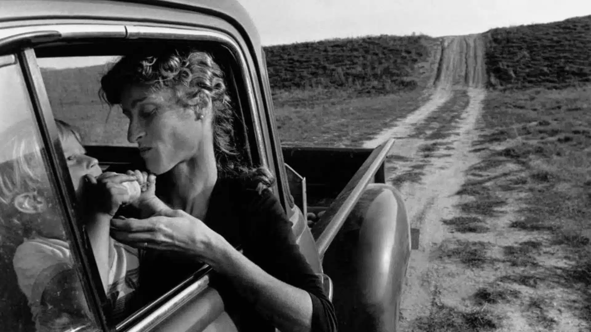 A woman leans into a pickup truck to kiss a child's hand as a dirt road climbs a grassy hill.
