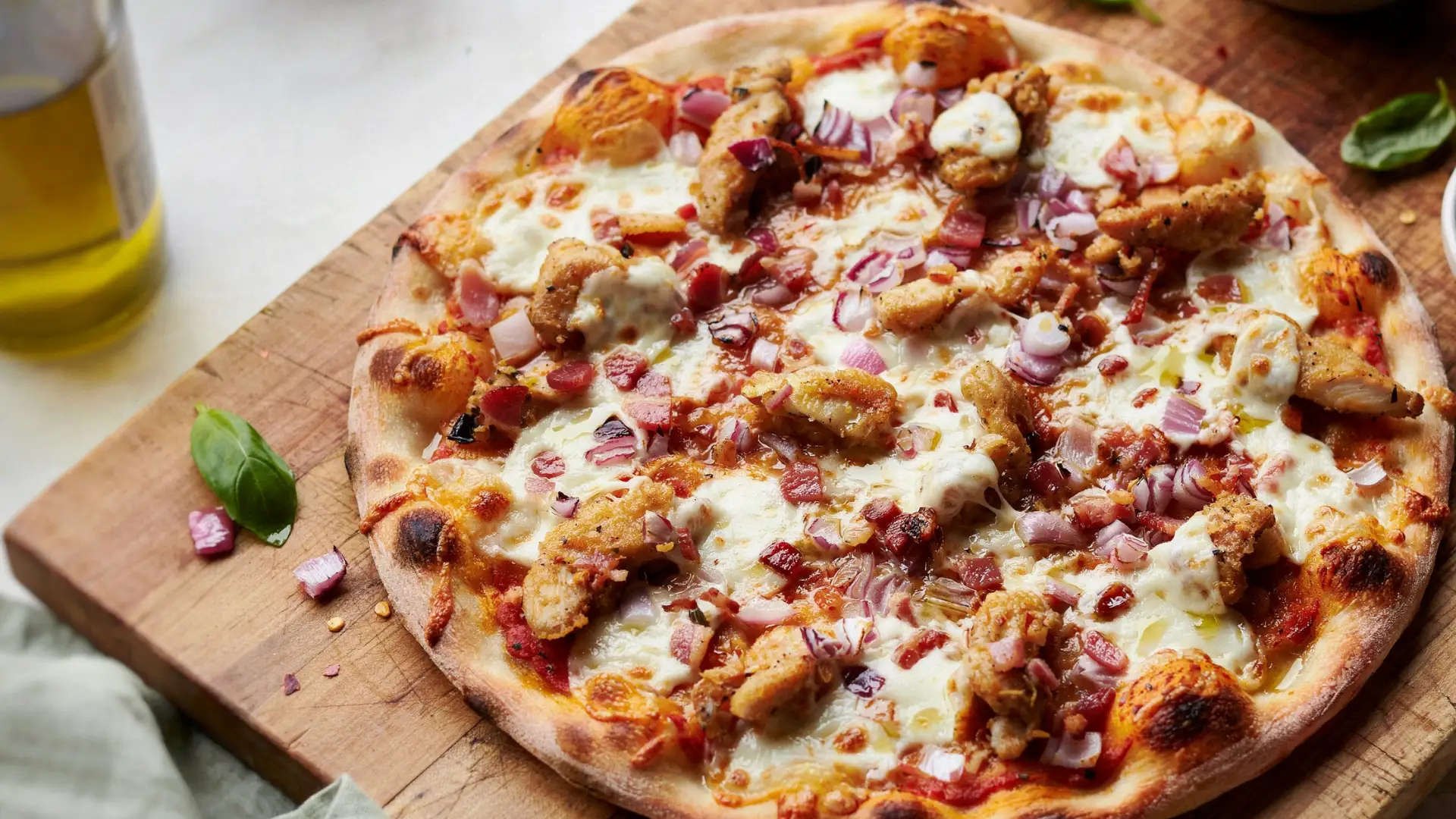 A wooden cutting board holds a pizza garnished with chicken, cheese, and onions.