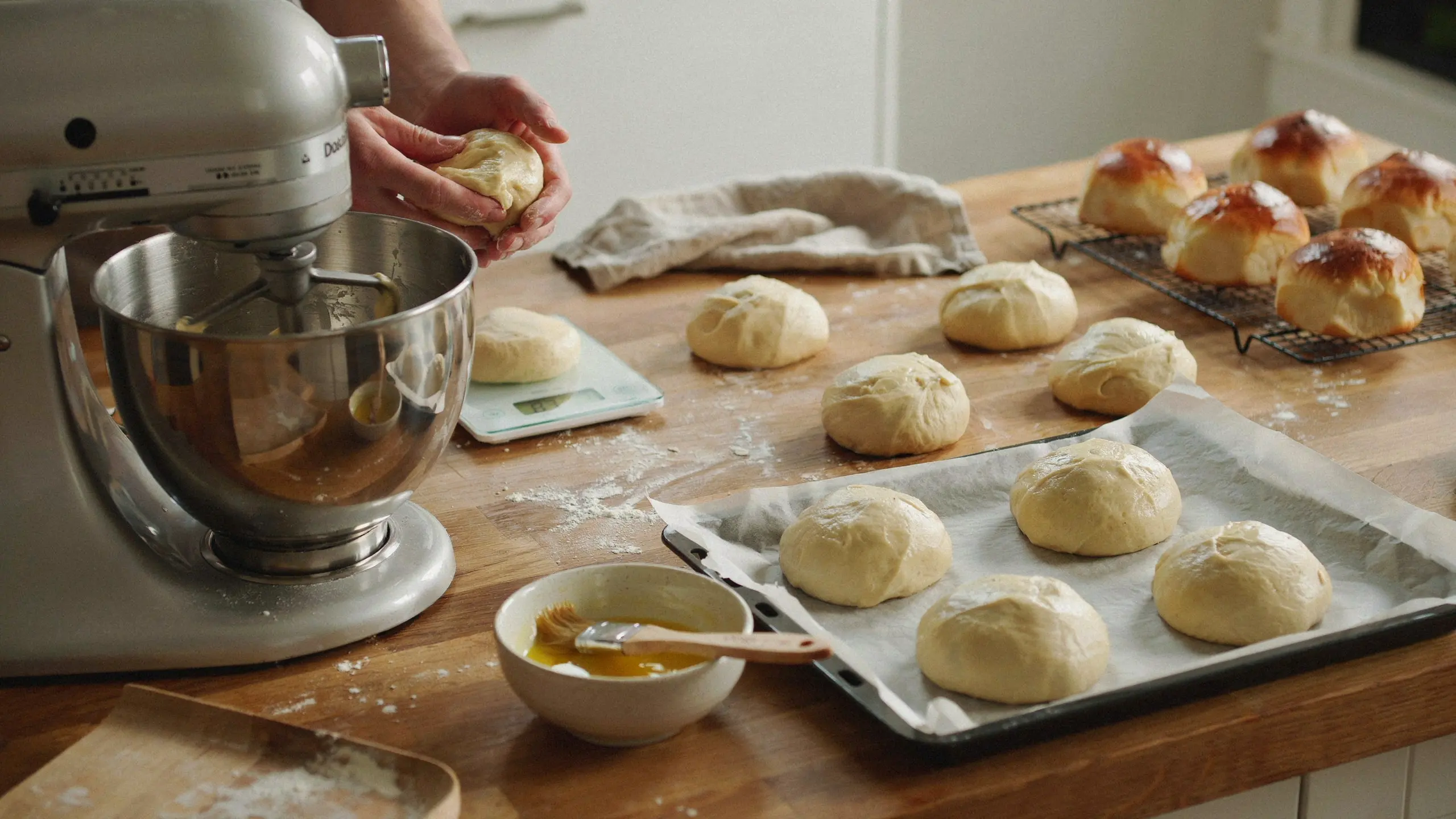 baker shaping brioche dough beside a stand mixer scale proofing tray and freshly baked buns on a cooling rack