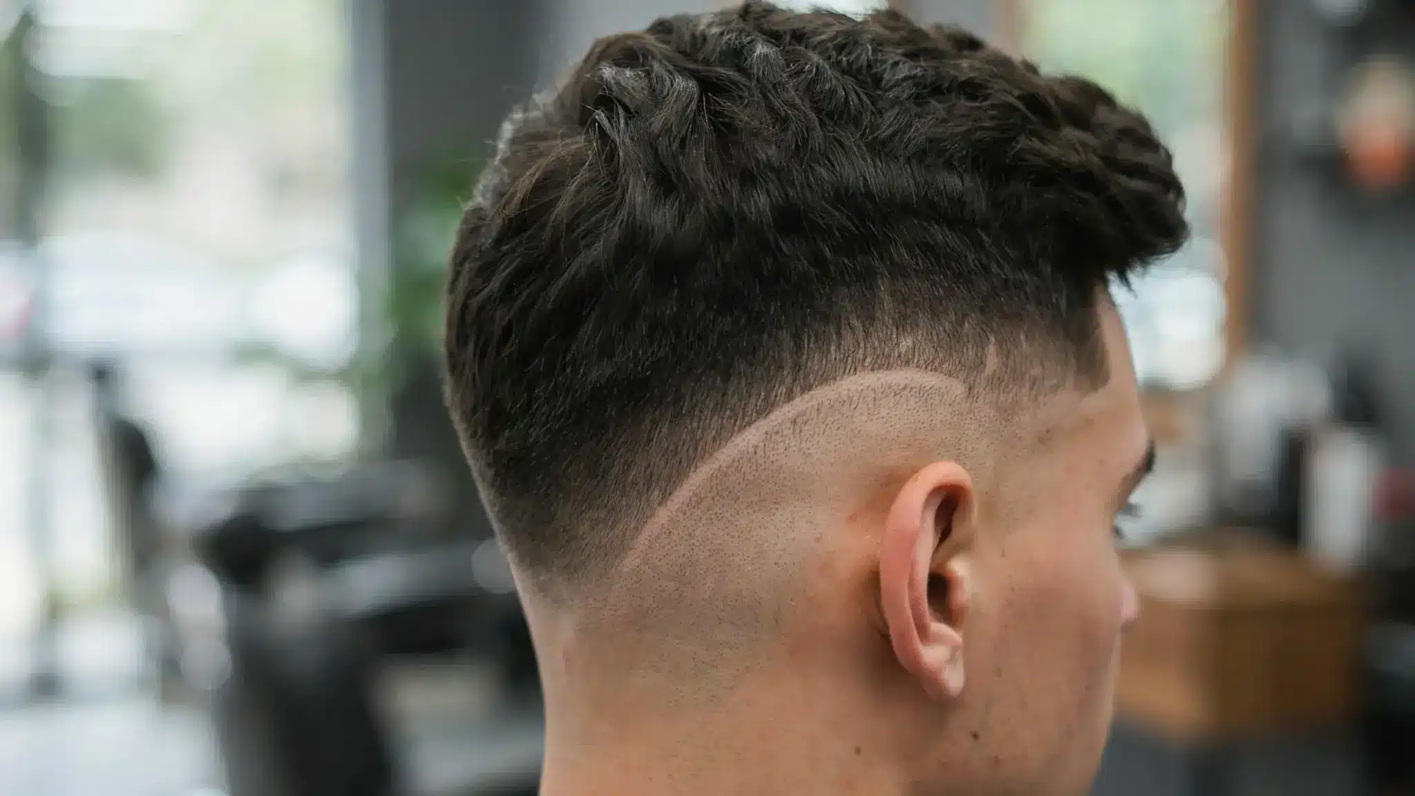 A man with high taper fade haircut is seated in a barber shop, surrounded by barber tools and equipment.