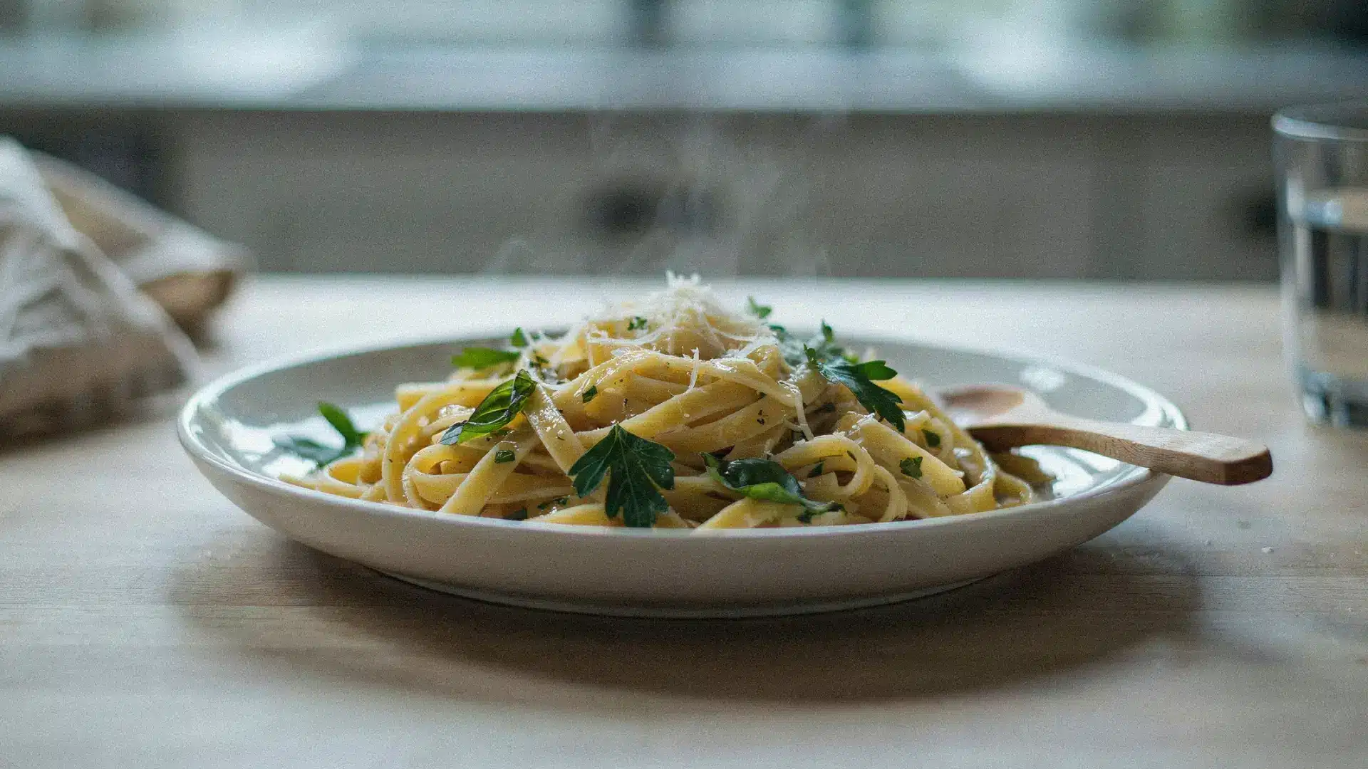 A plate of steaming hot fettuccine pasta garnished with fresh parsley and grated cheese sits on a wooden table, with a wooden spoon resting beside it.