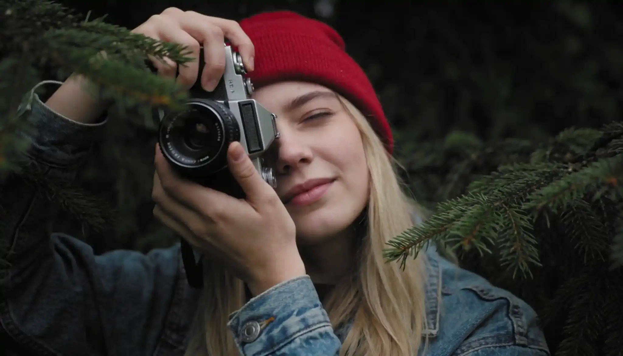 A young woman wearing a red beanie and denim jacket holds a vintage camera to her eye, surrounded by evergreen branches.