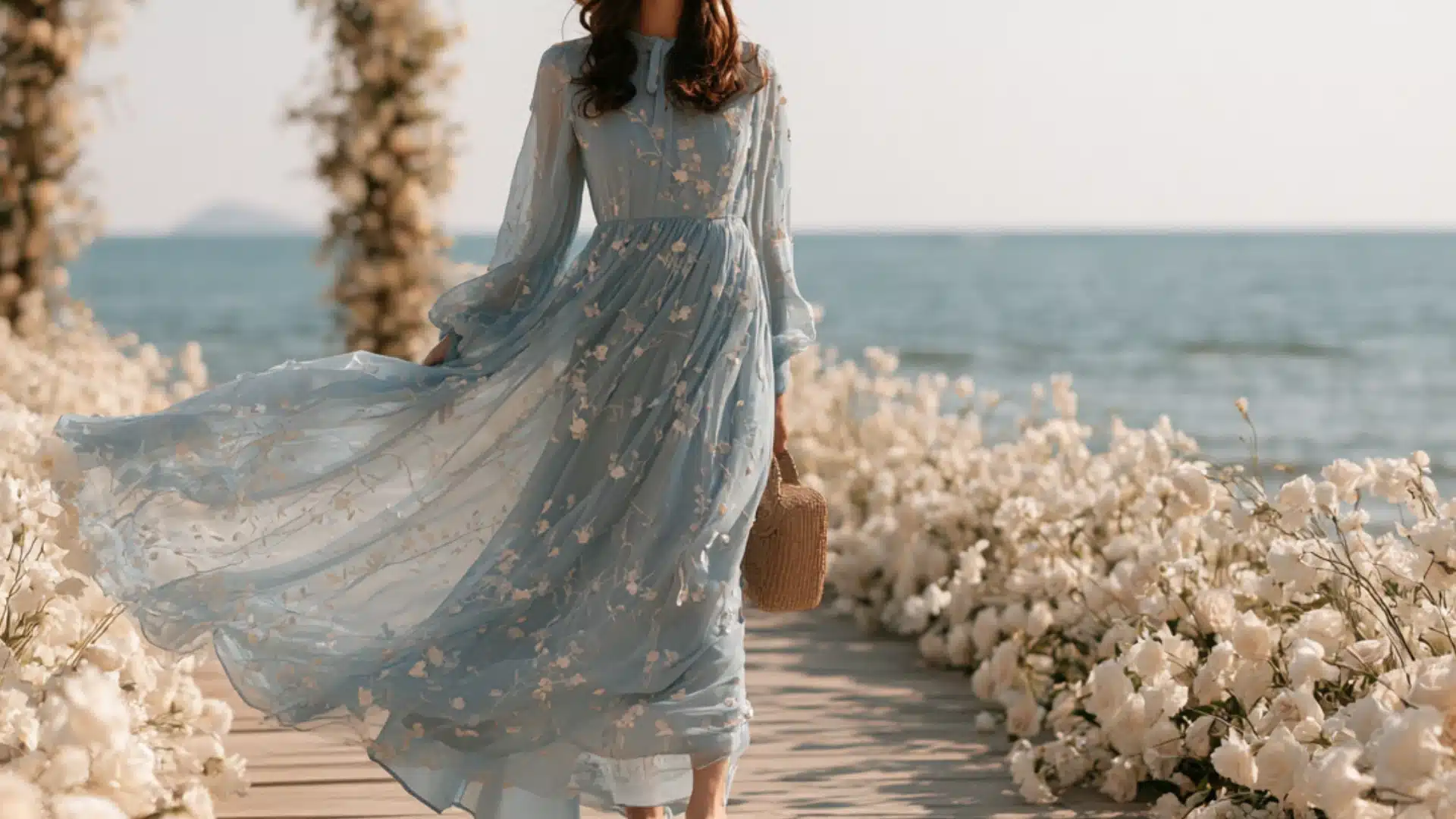 woman wearing a blue floral maxi dress with a woven bag walking through a beach wedding venue with white flowers