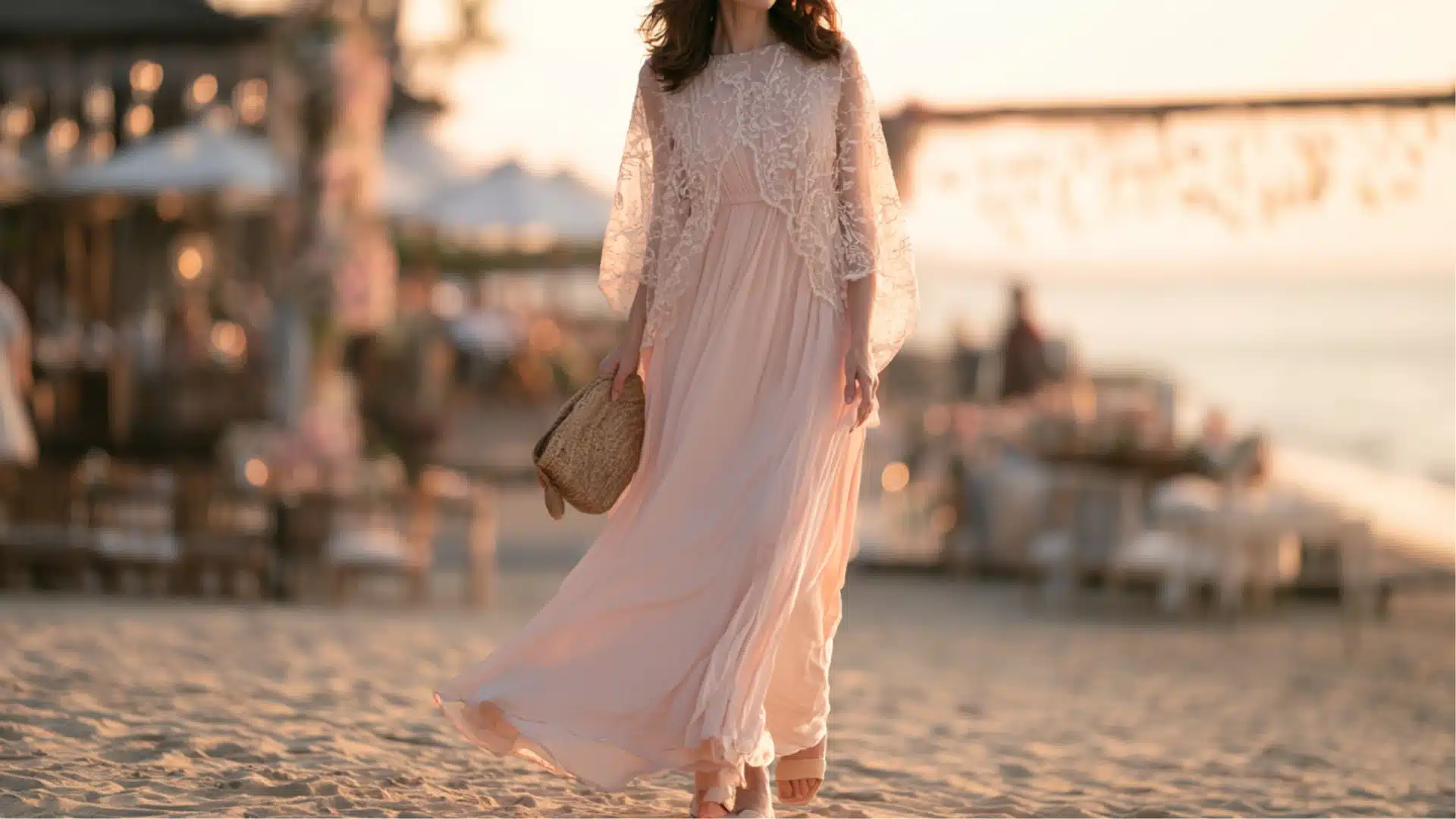 woman wearing a blush pink maxi dress with a sheer lace overlay walking along a beach wedding venue at sunset