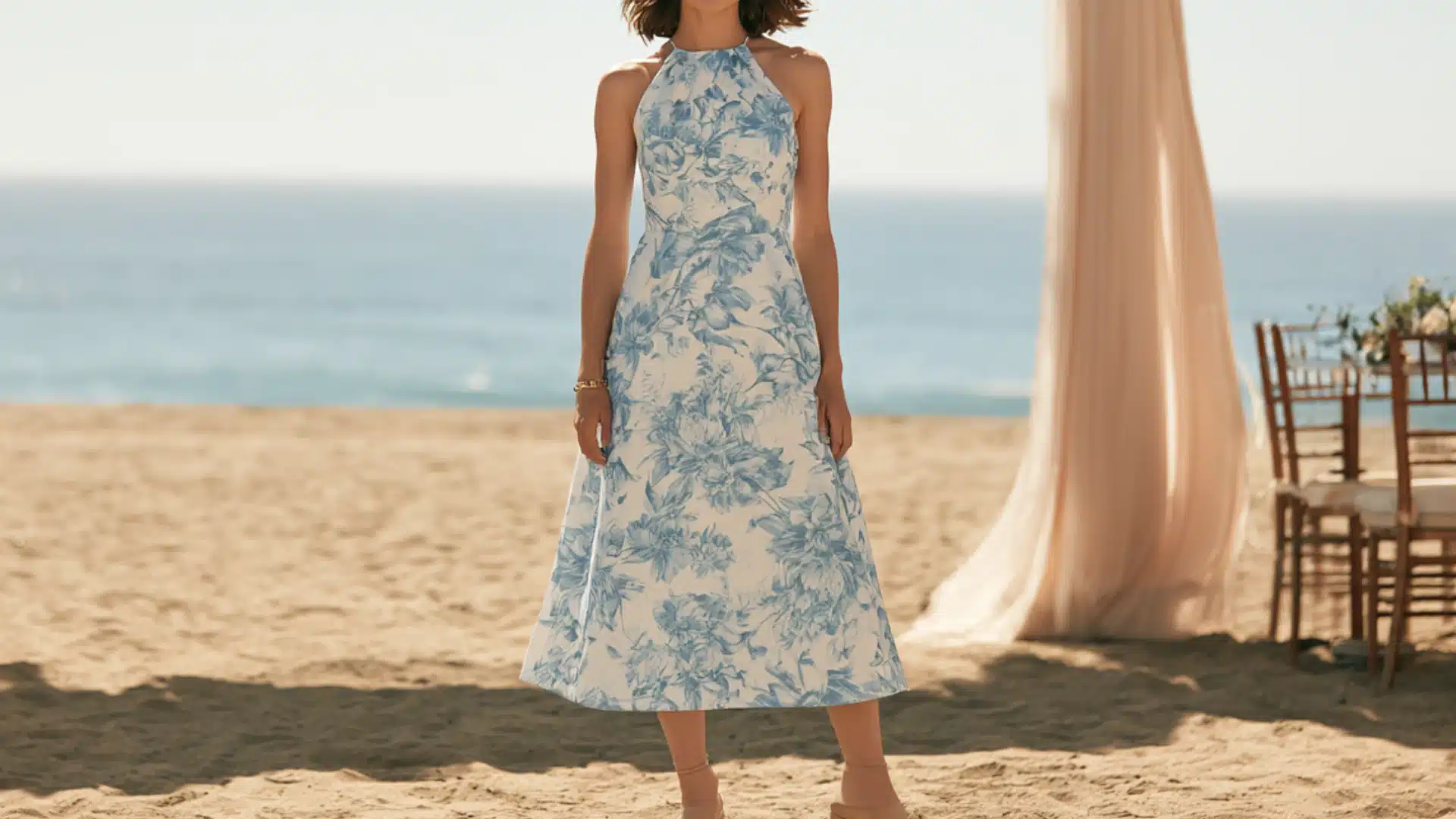woman wearing a light blue midi dress and block heel sandals walking through a beach wedding venue with white floral decorations